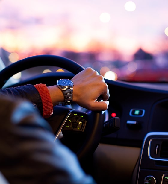 Main d'un conducteur avec une montre élégante sur le volant d'une voiture moderne au crépuscule. Lumières urbaines floues en fond.
