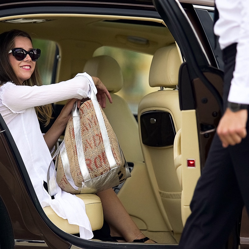 Femme souriante en lunettes assise dans une voiture de luxe beige. Elle tient un grand sac tissé argenté. Un chauffeur ouvre la porte.