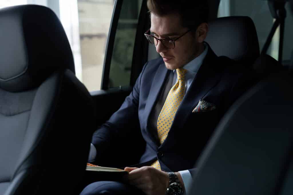 Homme d'affaires en costume sombre et cravate jaune, lunettes sur le nez, lisant un livre dans la banquette arrière d'une voiture, concentré.