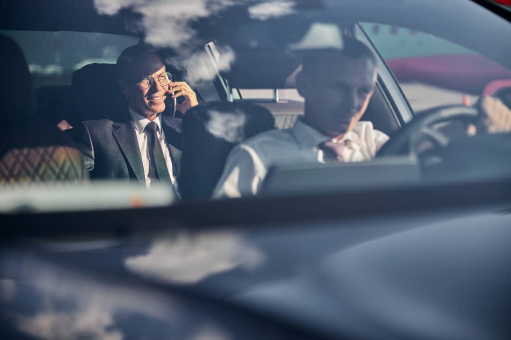 Homme d'affaires souriant en costume et lunettes, parlant au téléphone dans une voiture. Chauffeur flou au volant et reflets nuageux.