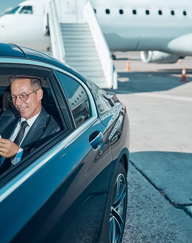 Homme souriant en costume et lunettes, chauffeured dans une voiture de luxe noire. Jet privé avec escaliers en arrière-plan à l'aéroport.