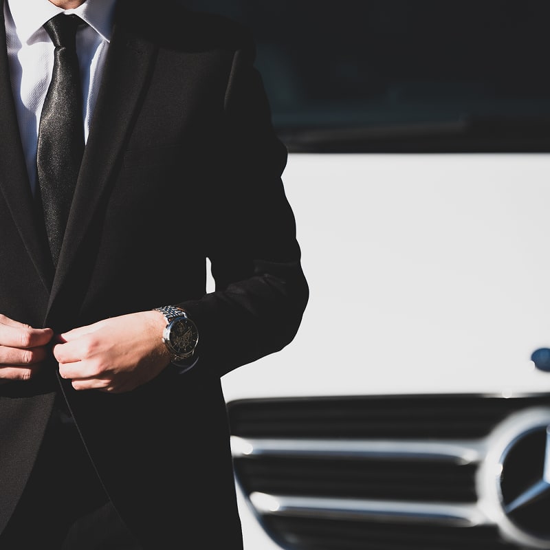 Homme élégant en costume noir, montre au poignet, ajustant sa veste devant une Mercedes-Benz blanche. Luxe et professionnalisme.