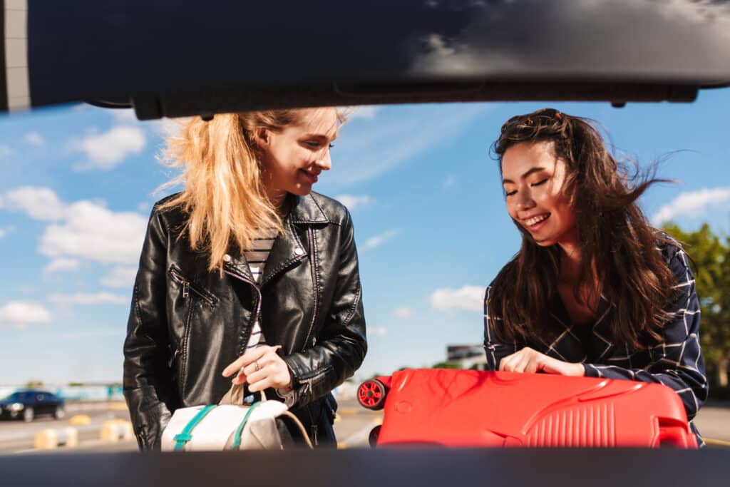 Deux amies souriantes chargent leurs bagages, dont une valise rouge vif, dans le coffre d'une voiture, sous un ciel bleu.