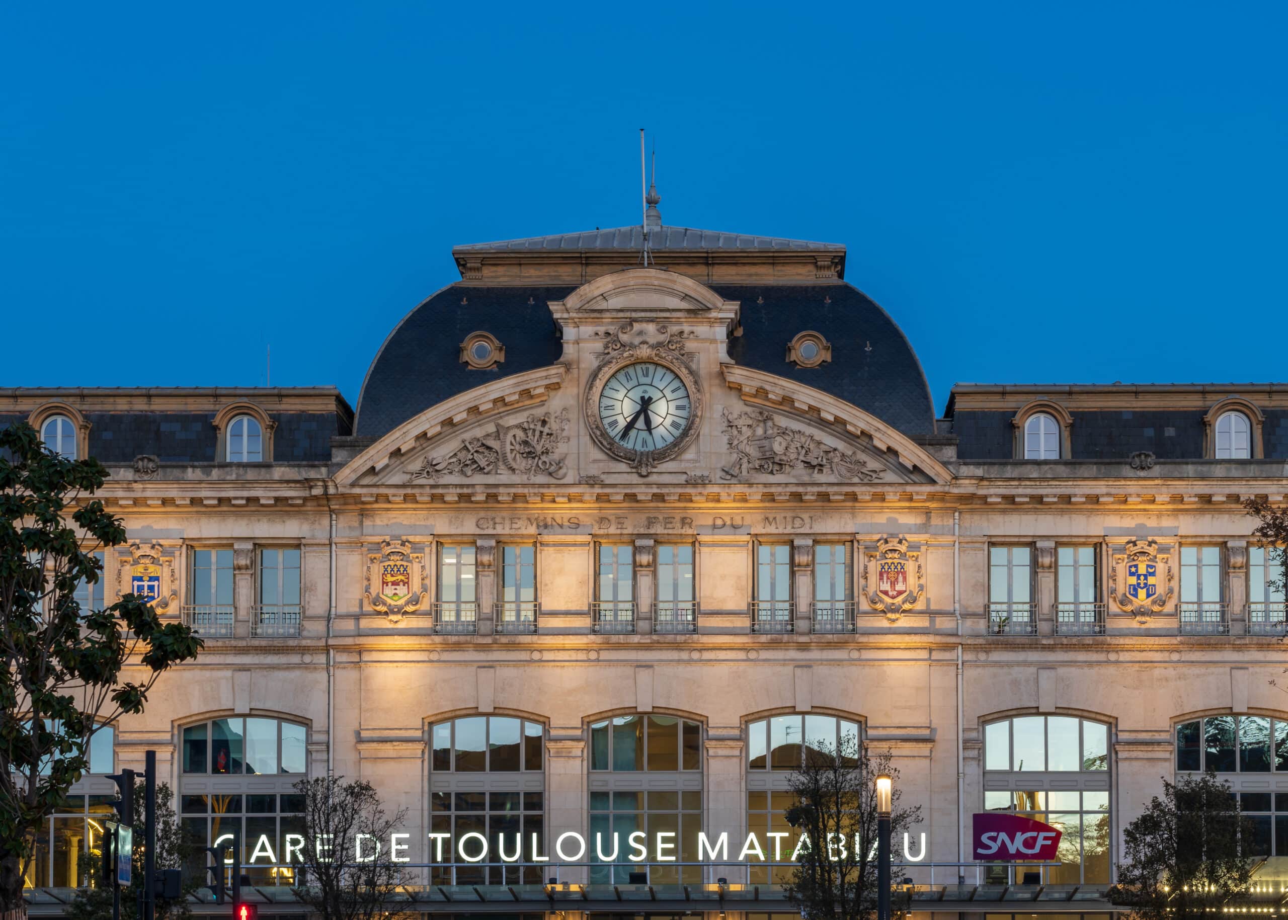 Façade illuminée de la Gare de Toulouse Matabiau au crépuscule. Horloge, sculptures ferroviaires, texte 'Chemins de fer du Midi', logo SNCF.