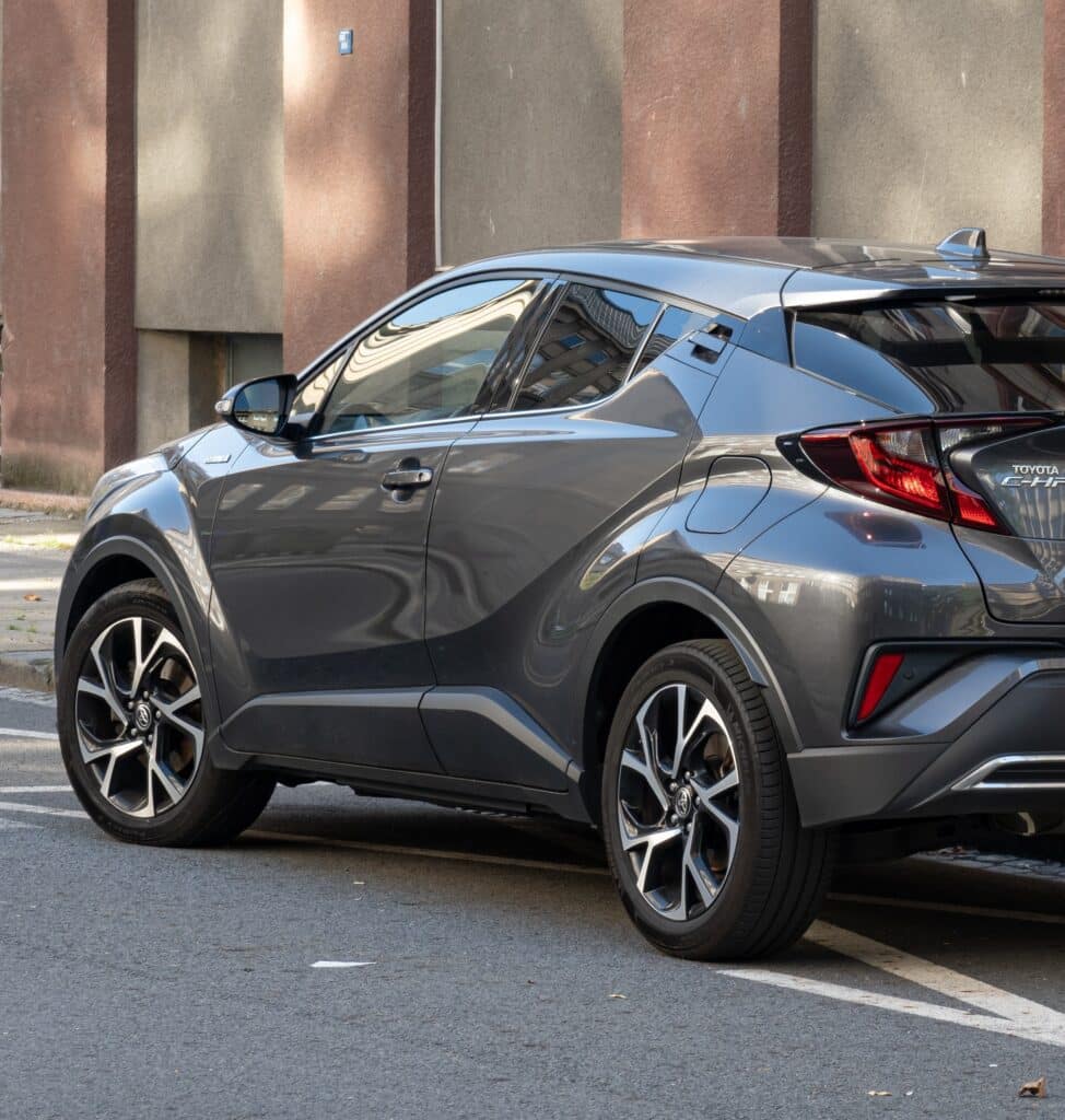 Toyota C-HR Hybride gris foncé, vue arrière-droite, garé sur l'asphalte avec réflexions urbaines. Texte: TOYOTA C-HR HYBRID.