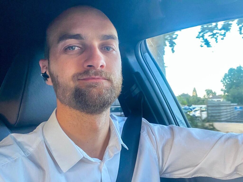 Homme élégant en chemise blanche, ceinture de sécurité, écouteur. Selfie dans une voiture, vue sur arbres et bâtiment par la fenêtre latérale.