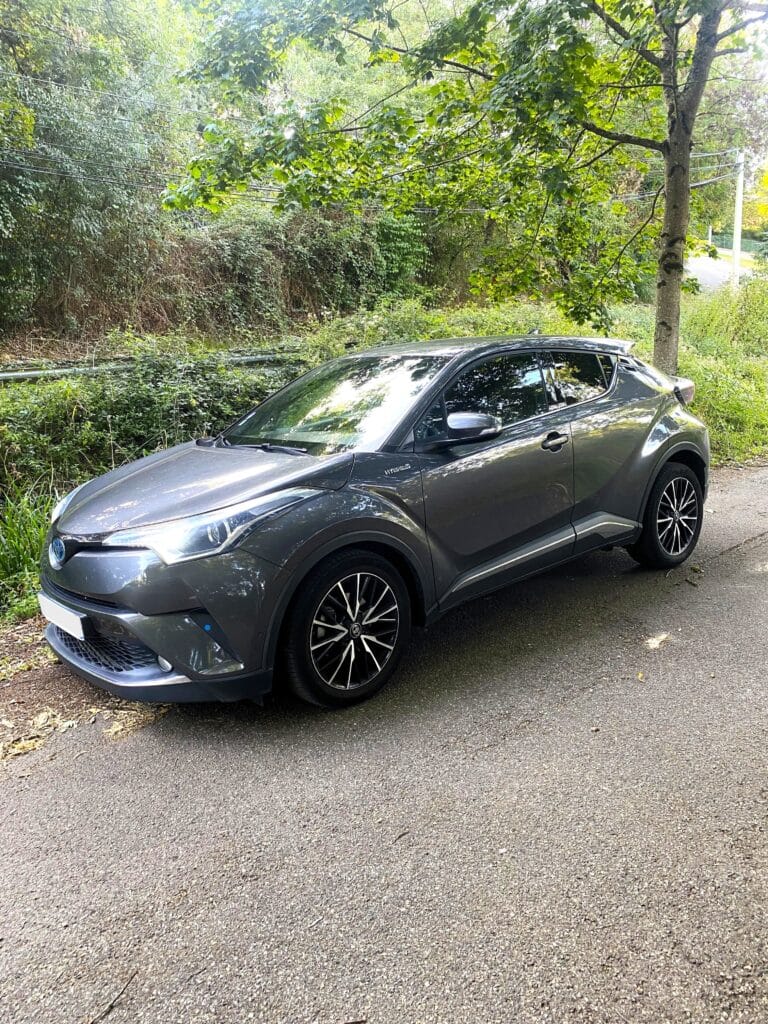 SUV Toyota C-HR hybride gris foncé garé sur une route asphaltée bordée de verdure dense et d'arbres. Badge 'HYBRID' visible.