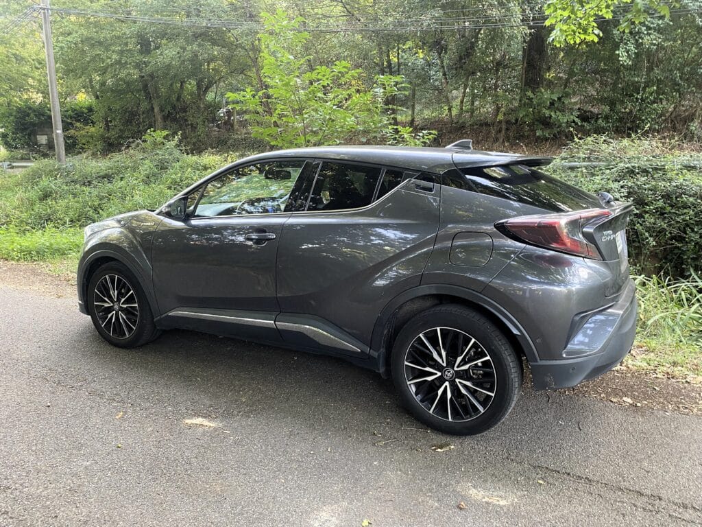 Toyota C-HR gris hybride, avec jantes bicolores, garée sur une route goudronnée devant une forêt dense et luxuriante.