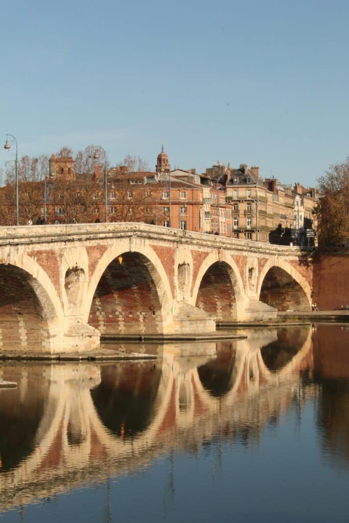Pont Neuf de Toulouse : Arches et reflets sur la Garonne Pont Neuf à Toulouse en briques rouges se reflétant dans la Garonne avec des bâtiments historiques en arrière-plan.